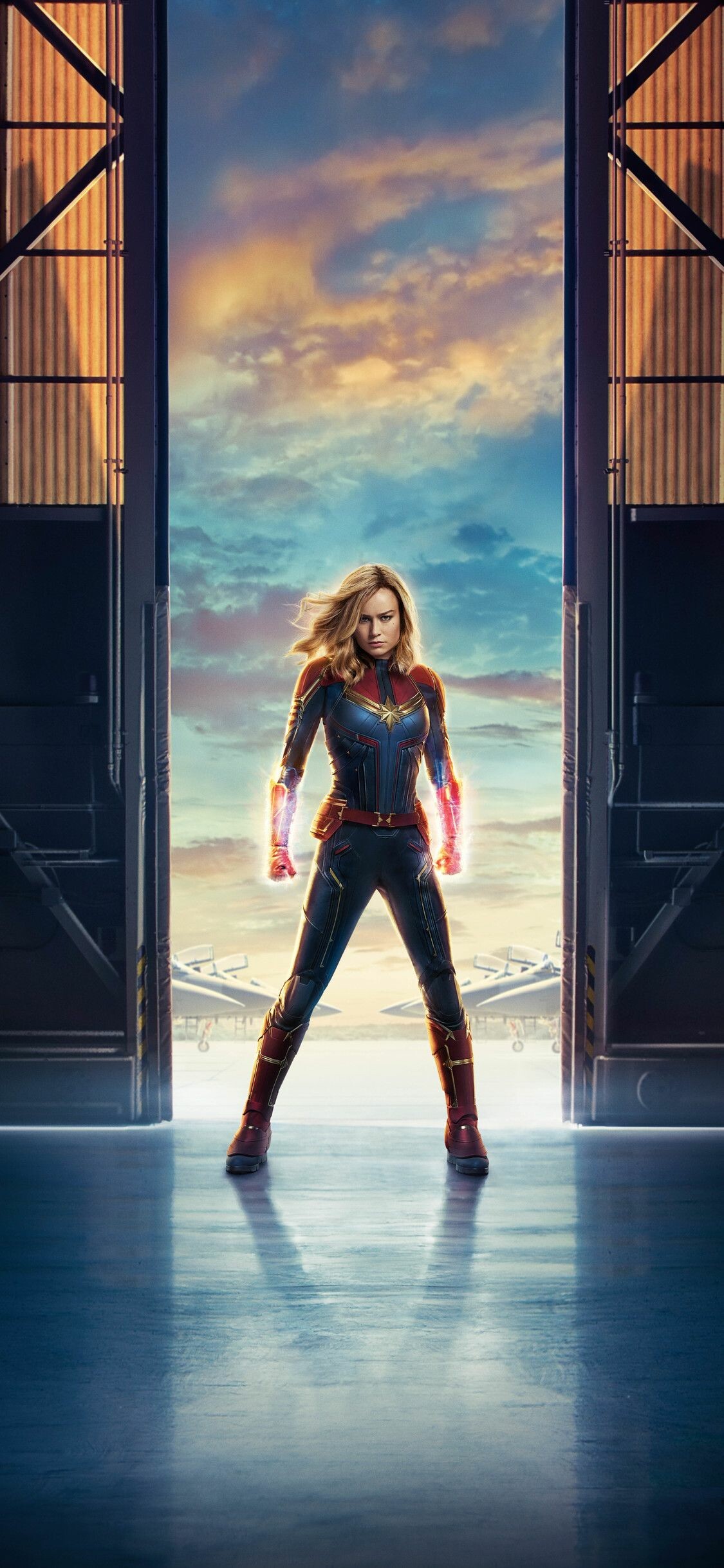 Brie Larson Captain Marvel, Hangar doorway silhouette, Glowing photon fists, Fighter jets tarmac — 1125×2436 HD wallpaper
